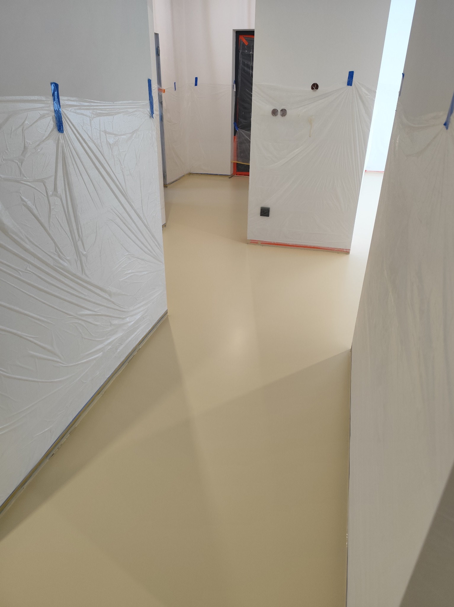 Beżowa, gładka podłoga w trakcie prac wykończeniowych. Ściany i drzwi zabezpieczone folią malarską. Widoczne taśmy i folie ochronne.