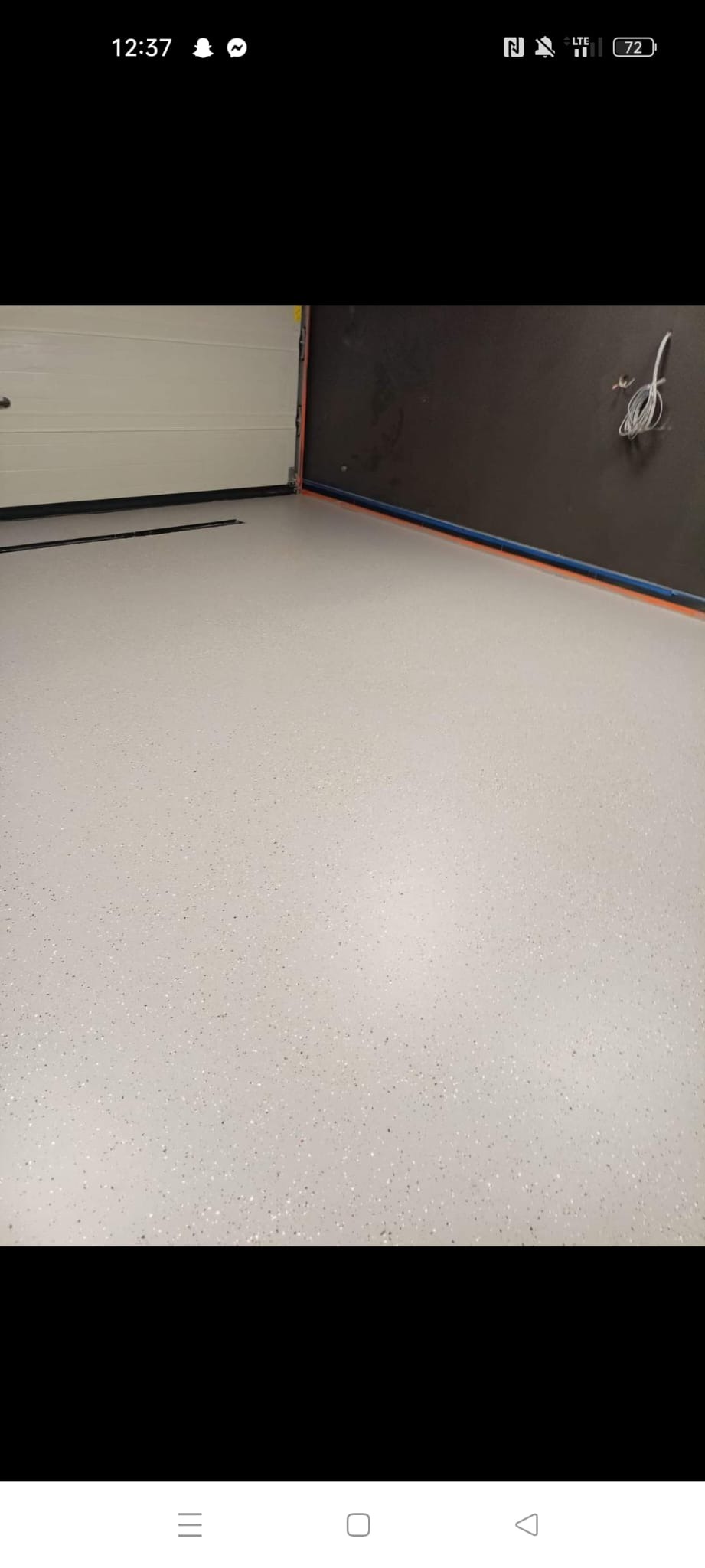 Jasnoszara posadzka żywiczna z drobnymi plamkami, widoczna brama garażowa i fragment ściany w ciemnym kolorze. Minimalistyczny design, gładka powierzchnia.