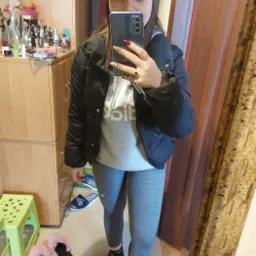 Osoba w czarnej kurtce i szarych legginsach robi selfie w lustrze, widoczny fragment pokoju z kosmetykami i butami.