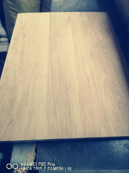 Obrabiany fragment płyty drewnianej o jasnym odcieniu i widocznym usłojeniu, leżący na niebieskim, prawdopodobnie metalowym podłożu. Widoczne ślady pyłu po obróbce.