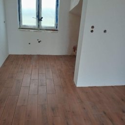 DOM MAR - Ułożona podłoga z brązowych płytek imitujących drewno w pokoju w trakcie remontu, okno zabezpieczone folią, ściany częściowo pomalowane na biało, widoczne otwory na gniazdka elektryczne.