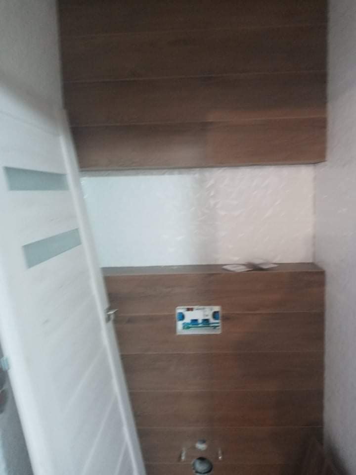 Ukończona łazienka z drewnopodobnymi płytkami na ścianach, widoczny fragment białych drzwi i puszka podtynkowego zestawu WC.