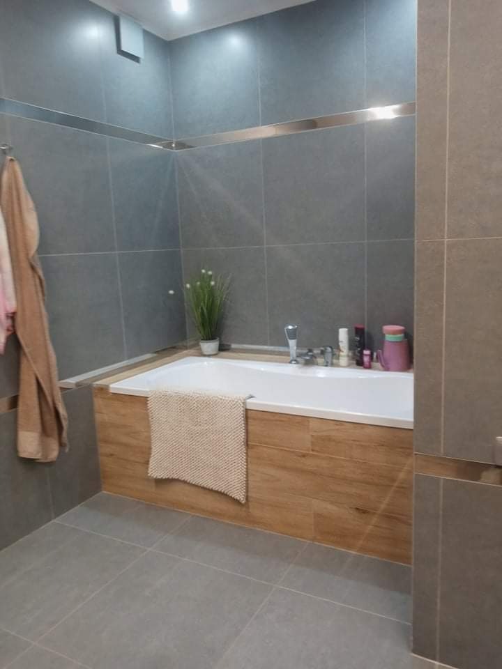 Nowoczesna łazienka z szarymi płytkami, obudową wanny imitującą drewno i dekoracyjną listwą ze stali nierdzewnej.