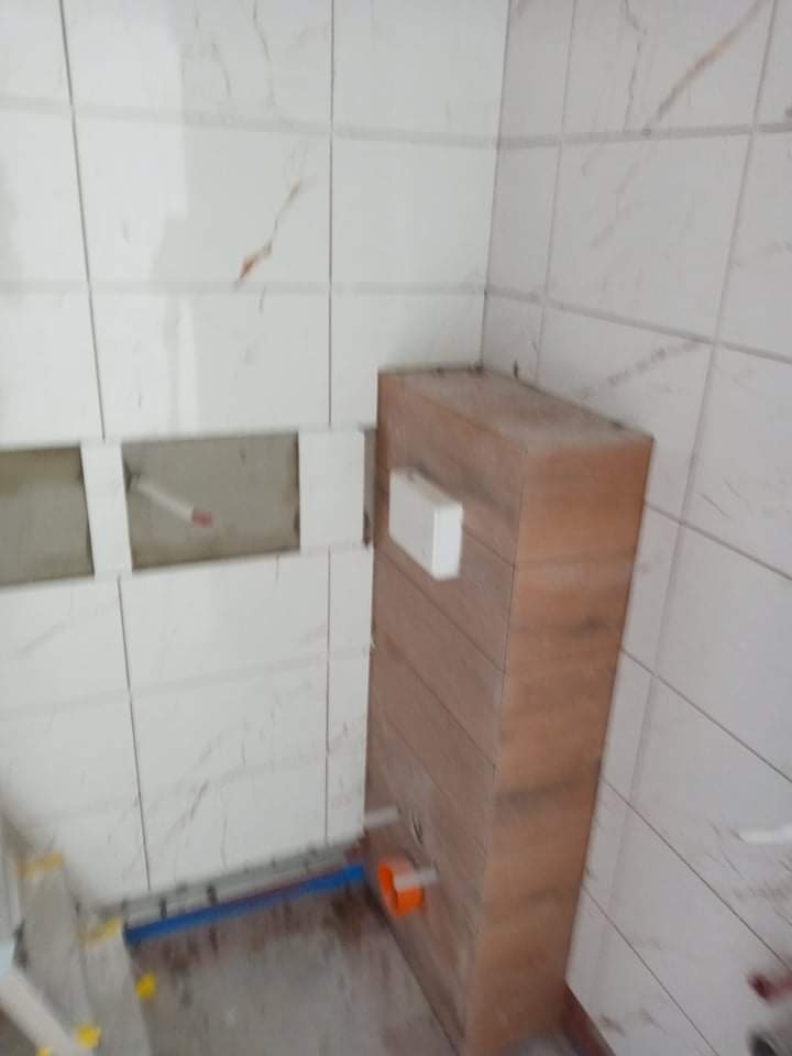 Niedokończona łazienka z częściowo położonymi białymi płytkami imitującymi marmur, widoczna zabudowa stelaża podtynkowego WC z drewnopodobną okładziną oraz fragment instalacji wodno-kanalizacyjnej.