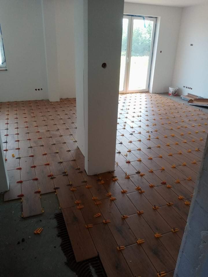 Układanie drewnopodobnych płytek podłogowych w jasnym wnętrzu, z użyciem pomarańczowych klipsów poziomujących, widoczna częściowo ułożona podłoga i kolumna.