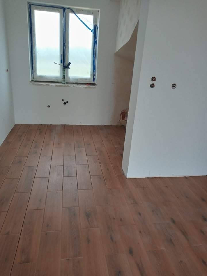 Ułożona podłoga z brązowych płytek imitujących drewno w pokoju w trakcie remontu, okno zabezpieczone folią, ściany częściowo pomalowane na biało, widoczne otwory na gniazdka elektryczne.