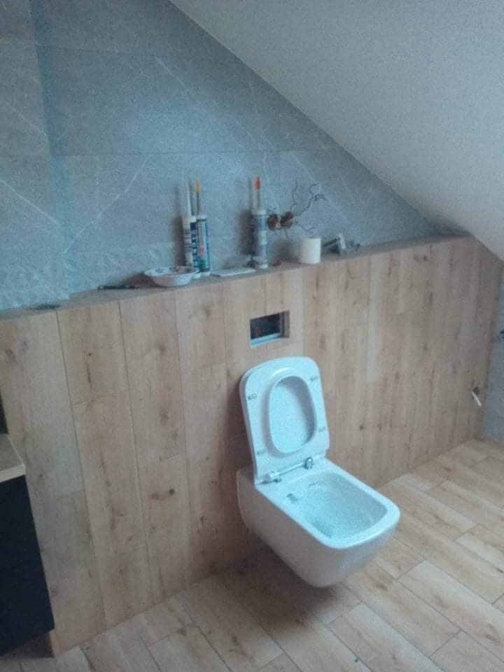 Podwieszana toaleta z deską w kolorze białym zamontowana na ścianie wyłożonej panelami imitującymi drewno, widoczne akcesoria montażowe i silikony.