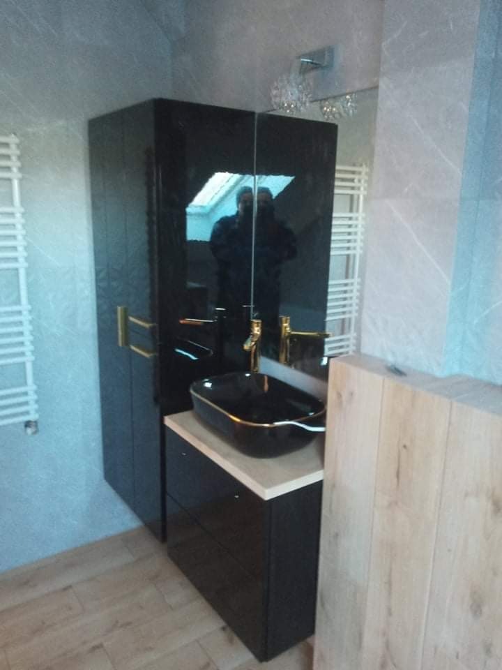 Nowoczesna łazienka w Żninie z czarną szafką pod umywalkę, złotą armaturą i lustrem, które odbija okno dachowe.