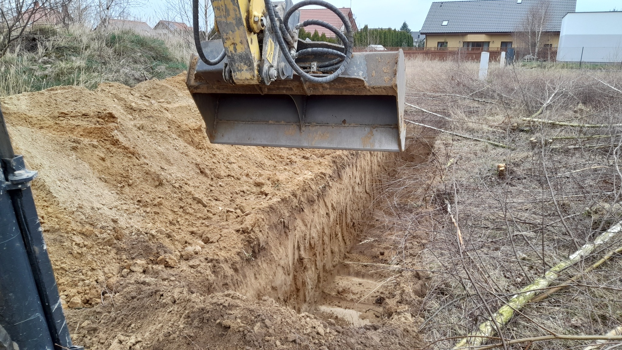 Żółta koparka z łyżką głęboko w ziemi, wykopany wąski rów w piaszczystej glebie, w tle zabudowania mieszkalne i suche krzewy.