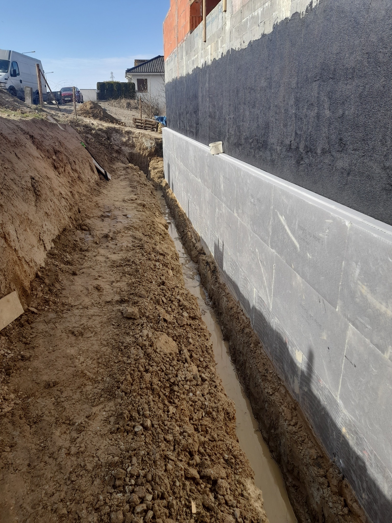Fundament budynku zabezpieczony izolacją pionową, widoczny wykop wzdłuż ściany fundamentowej wypełniony wodą, ziemia.