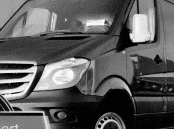 Częściowy widok przodu czarnego busa z widoczną tabliczką za przednią szybą, zaparkowanego w pomieszczeniu.