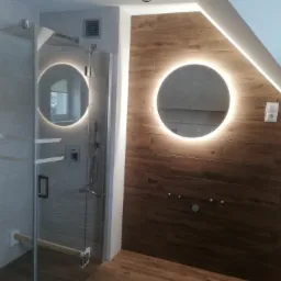 Nowoczesna łazienka z kabiną prysznicową ze szklanymi drzwiami, oświetlonymi okrągłymi lustrami na ścianach wyłożonych płytkami imitującymi drewno i szarymi płytkami strukturalnymi.