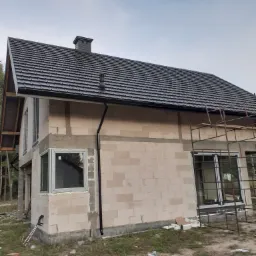 Dom w trakcie budowy z nowym, ciemnoszarym dachem z dachówki ceramicznej, widoczne rynny, komin i rusztowanie przy elewacji z betonu komórkowego.