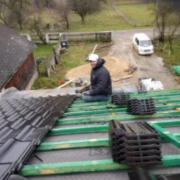 Dekarz w kurtce i czapce siedzi na nowo krytym dachu z czarnej dachówki, w tle widoczny samochód dostawczy i krajobraz wiejski.