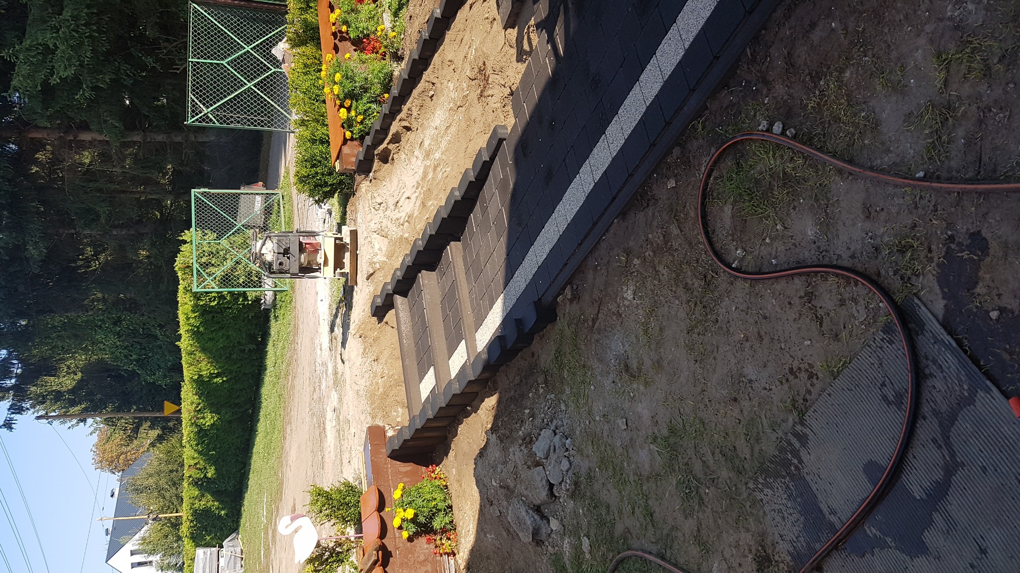 Przygotowany teren pod ścieżkę z ułożonymi obrzeżami z kostki i częściowo ułożoną kostką brukową, widoczna zagęszczarka w tle.