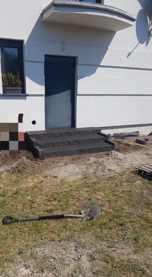 Ułożone schody wejściowe z ciemnej kostki granitowej przed domem w Wieleniu, widoczna łopata na trawniku i częściowo wykończona elewacja.