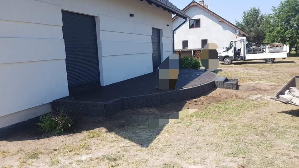 Wykończony taras z ciemnej kostki brukowej przy białym domu z szarą bramą garażową, widoczne schodki z kostki i samochód dostawczy w tle.