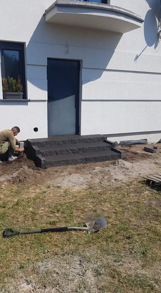 Montaż granitowych schodów wejściowych do domu jednorodzinnego; pracownik w stroju roboczym układa ostatni element, widoczna łopata na trawie.