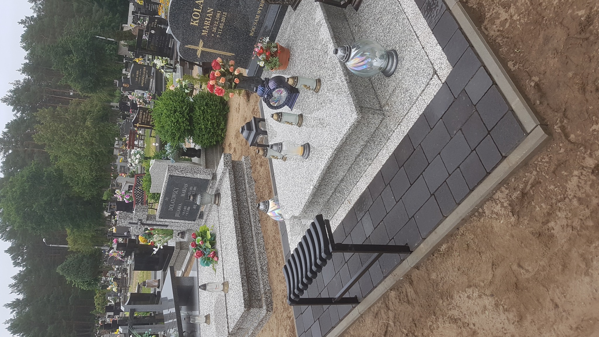 Częściowo wyłożona kostką granitową przestrzeń wokół nagrobka na cmentarzu, widoczne świeże ślady ziemi i czarne ławki, w tle inne groby i drzewa.