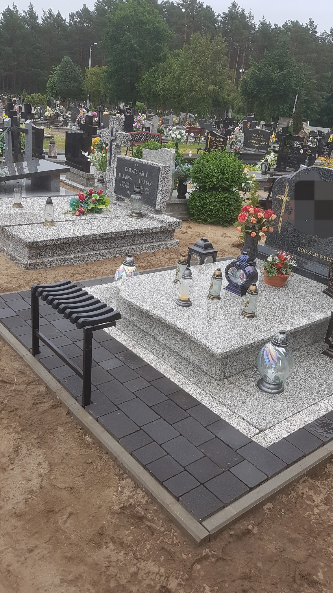 Ciemnoszara kostka brukowa ułożona wokół granitowego nagrobka na cmentarzu, z czarną metalową ławeczką i licznymi lampionami oraz kwiatami.