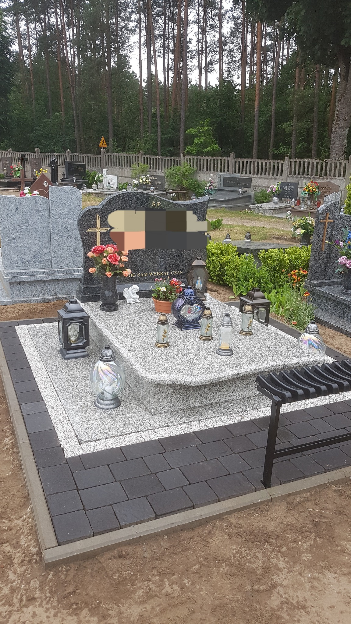 Granitowy nagrobek z ciemną kostką brukową wokół, ozdobiony kwiatami, zniczami i figurką anioła na cmentarzu.