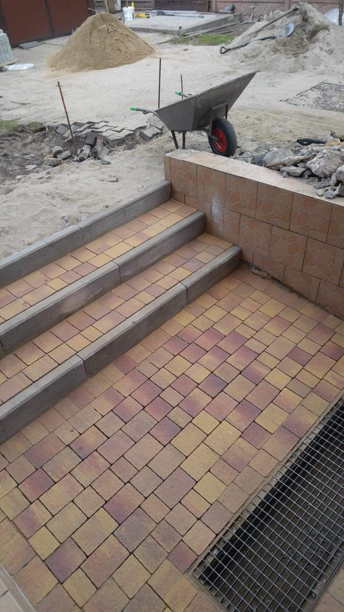 Schody wyłożone kostką brukową w odcieniach żółci i czerwieni, z szarymi obrzeżami, prowadzące do poziomu gruntu z kratką ściekową; w tle sterta piasku i taczka.