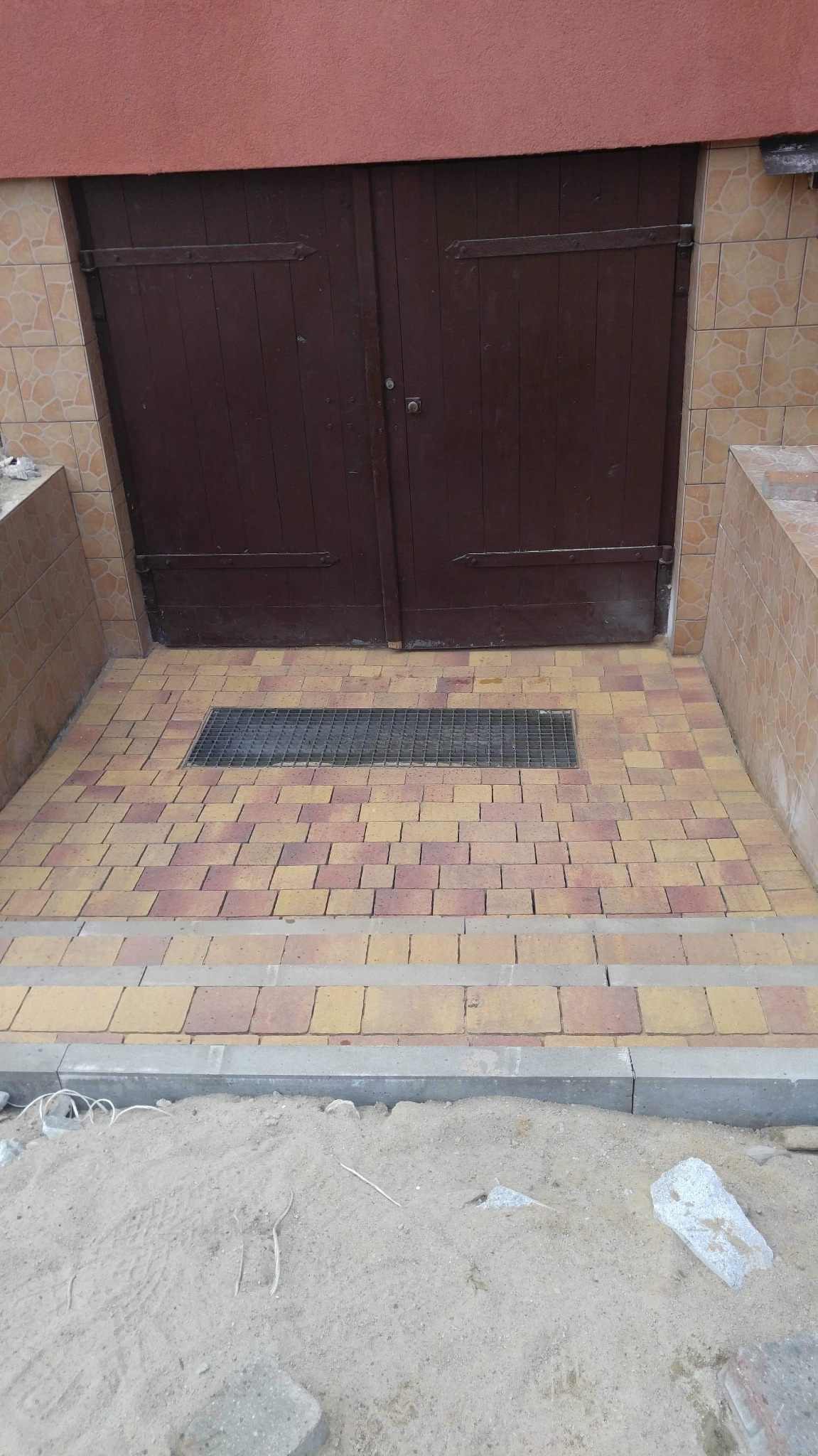 Ułożona kostka brukowa w odcieniach żółci i czerwieni przed brązowymi drzwiami z metalową kratką odpływową, widoczne schody i piasek z resztkami materiałów budowlanych.