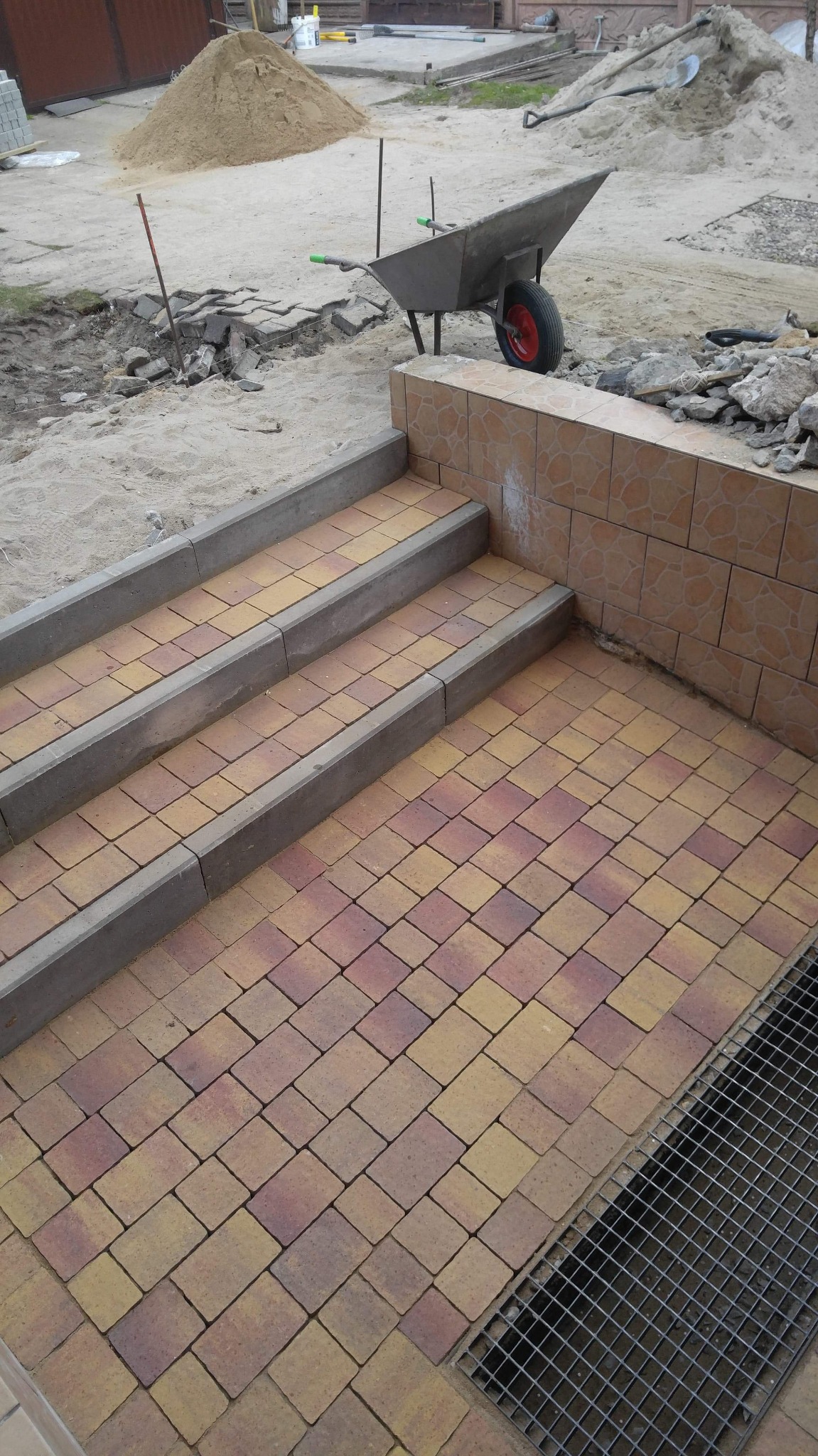 Ułożone schody z kostki brukowej w odcieniach żółci i czerwieni, z betonowymi obrzeżami, obok wózek budowlany z piaskiem w tle.