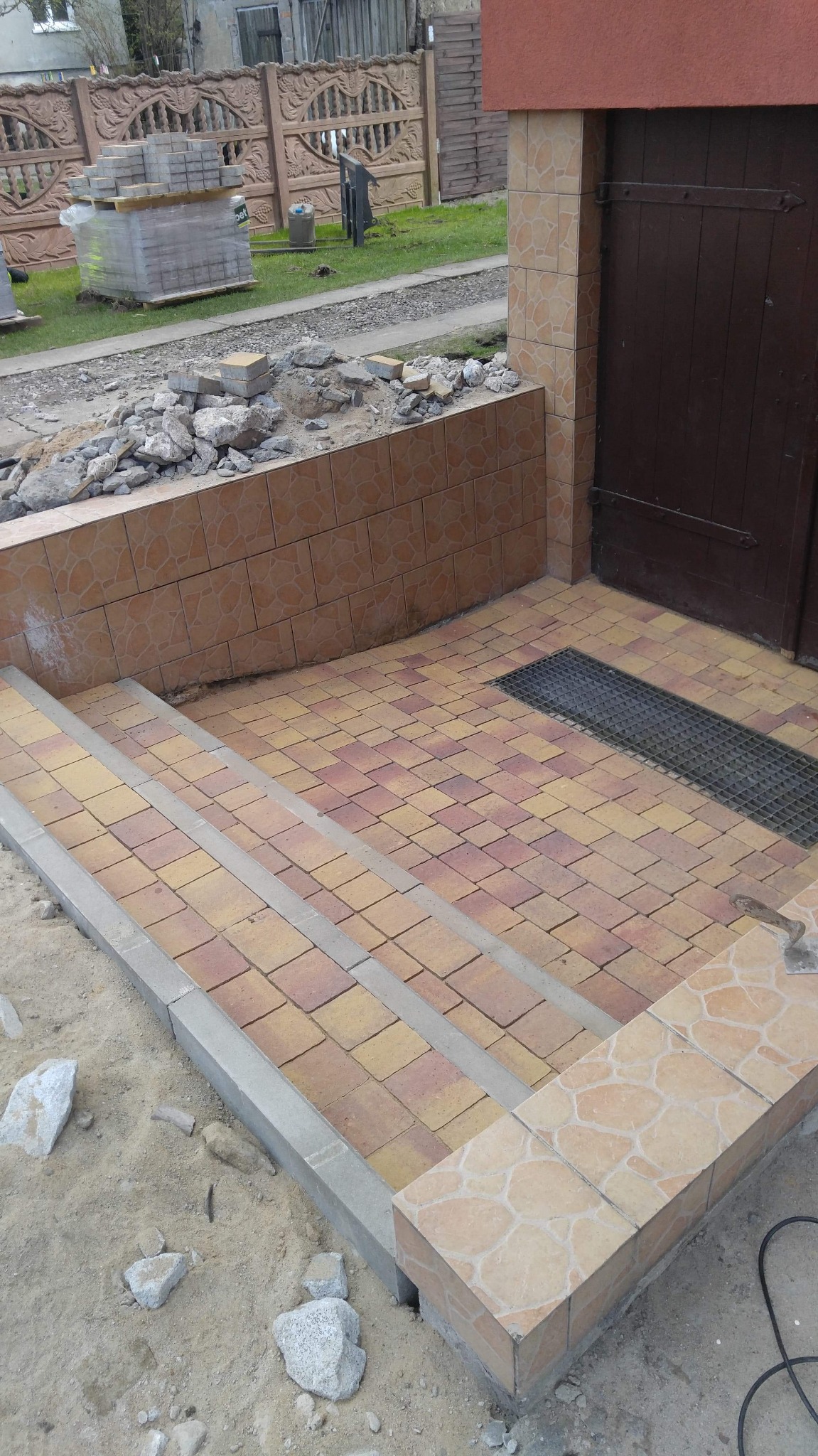 Schody wejściowe wyłożone kostką brukową w odcieniach żółci i czerwieni, z szarą obwódką, prowadzące do brązowych drzwi; widoczna kratka odpływowa i fragment muru z dekoracyjnymi płytkami.