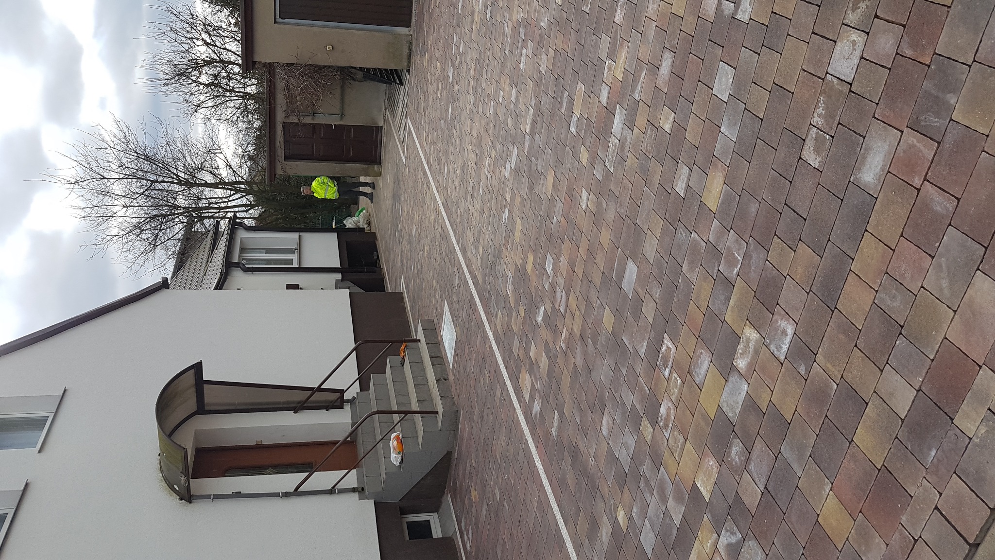 Ułożona kostka brukowa w odcieniach brązu i beżu przed budynkiem z białymi ścianami i szarymi schodami, widoczna osoba w jaskrawej kamizelce odblaskowej.