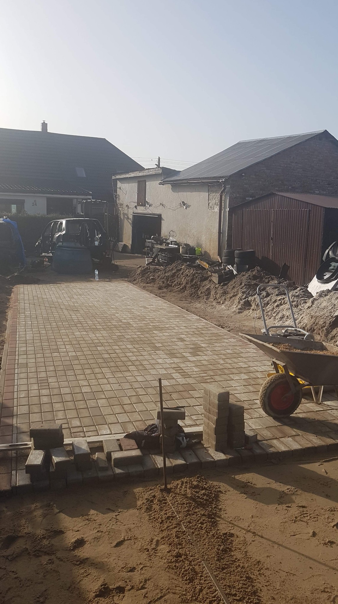 Prostokątny podjazd z kostki brukowej w trakcie układania, widoczny wózek budowlany z piaskiem, zaparkowany samochód w tle.