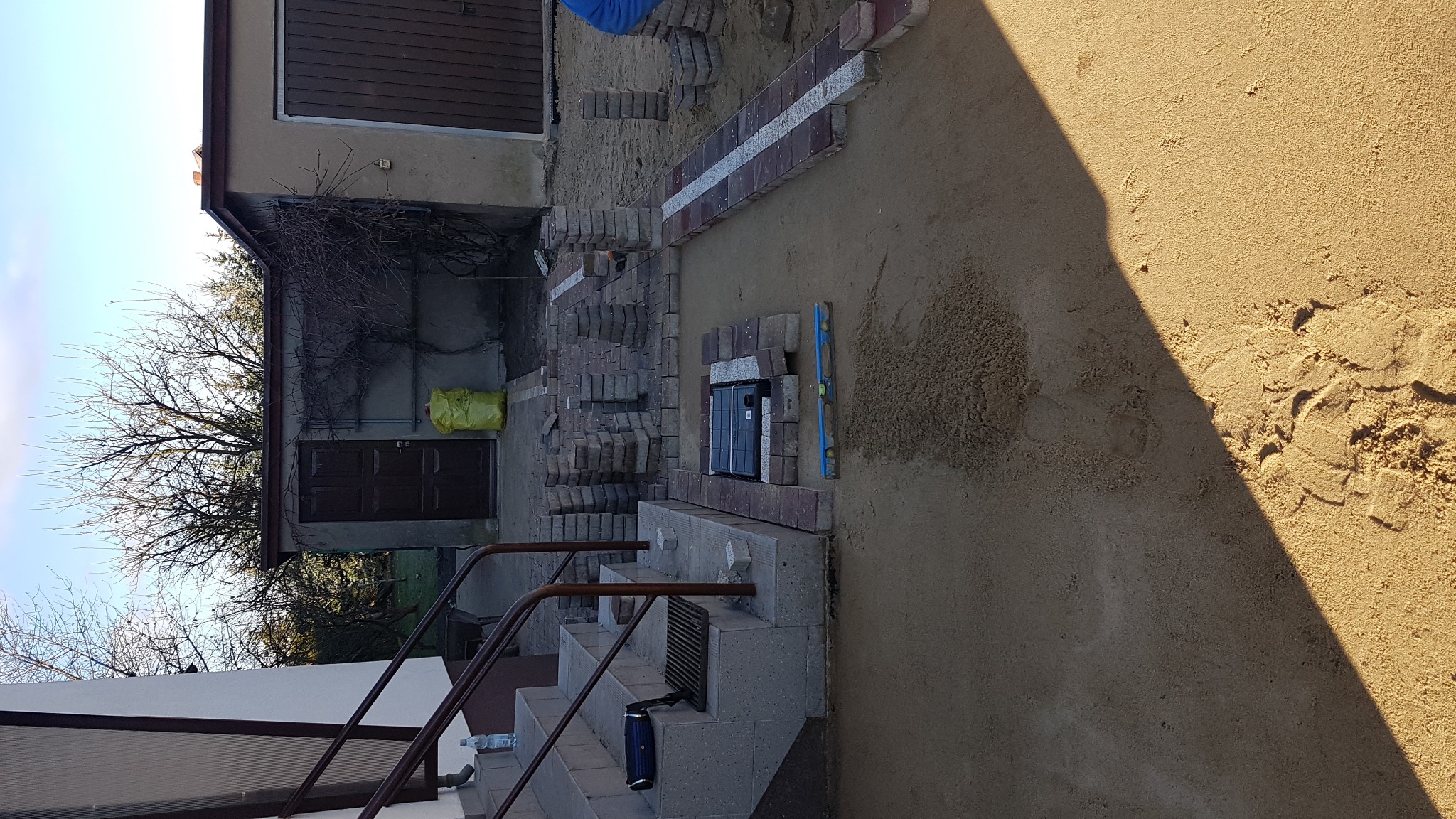 Prace brukarskie w toku: ułożona część ścieżki z czerwono-szarych kostek, poziomica i smartfon na kostce, piasek, w tle budynek gospodarczy i schody.