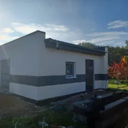 Nowoczesny, parterowy budynek z jasną elewacją, poziomym pasem w kolorze grafitowym, grafitowymi drzwiami i bramą garażową, widok z zewnątrz, w tle drzewa i błękitne niebo z promieniami słońca.