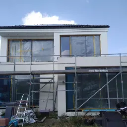 Dwukondygnacyjny dom w trakcie tynkowania elewacji, okna zabezpieczone folią, widoczne rusztowania i drabina, w tle błękitne niebo.