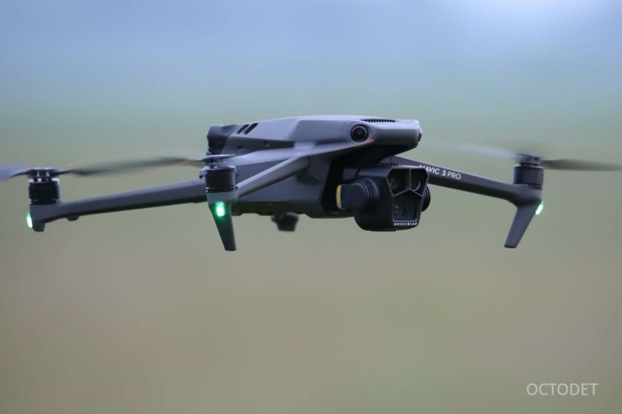 Szary dron Mavic 3 Pro w locie na tle rozmytego, zielono-szarego nieba. Widoczne zielone światła pozycyjne i kamera Hasselblad.