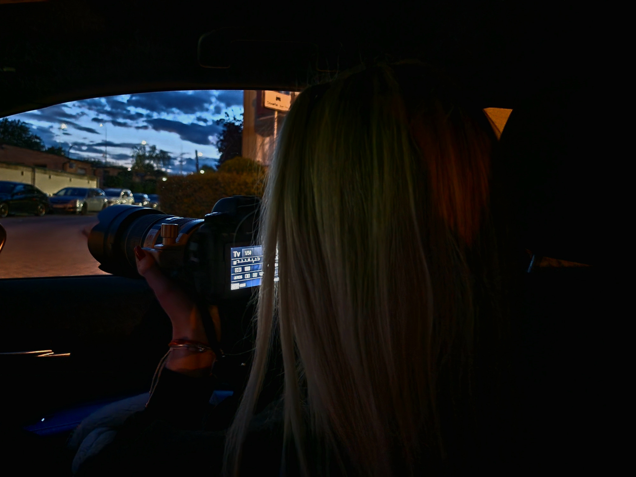 Kobieta z długimi blond włosami siedzi w samochodzie i robi zdjęcia aparatem z dużym obiektywem, obserwując otoczenie na zewnątrz w nocy.