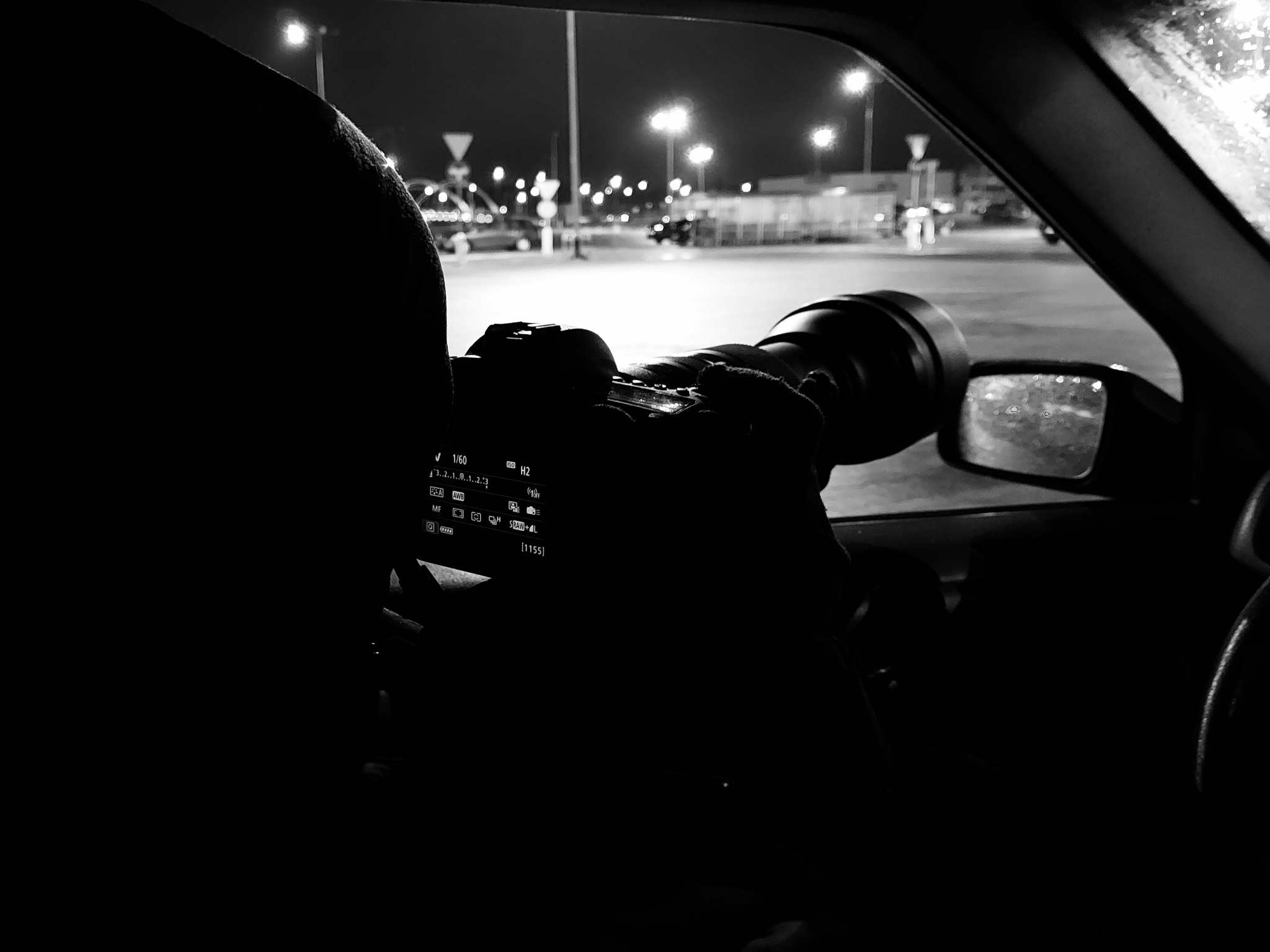 Osoba z wnętrza samochodu, z dużym obiektywem, robi zdjęcie nocnego parkingu z widocznymi światłami ulicznymi i kroplami deszczu na szybie.