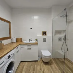 Nowoczesna łazienka z drewnianymi akcentami: blat z jasnego drewna, podłoga imitująca drewno, białe płytki ścienne, kabina prysznicowa ze szklaną ścianką, wisząca toaleta i pralka Bosch Maxx 7