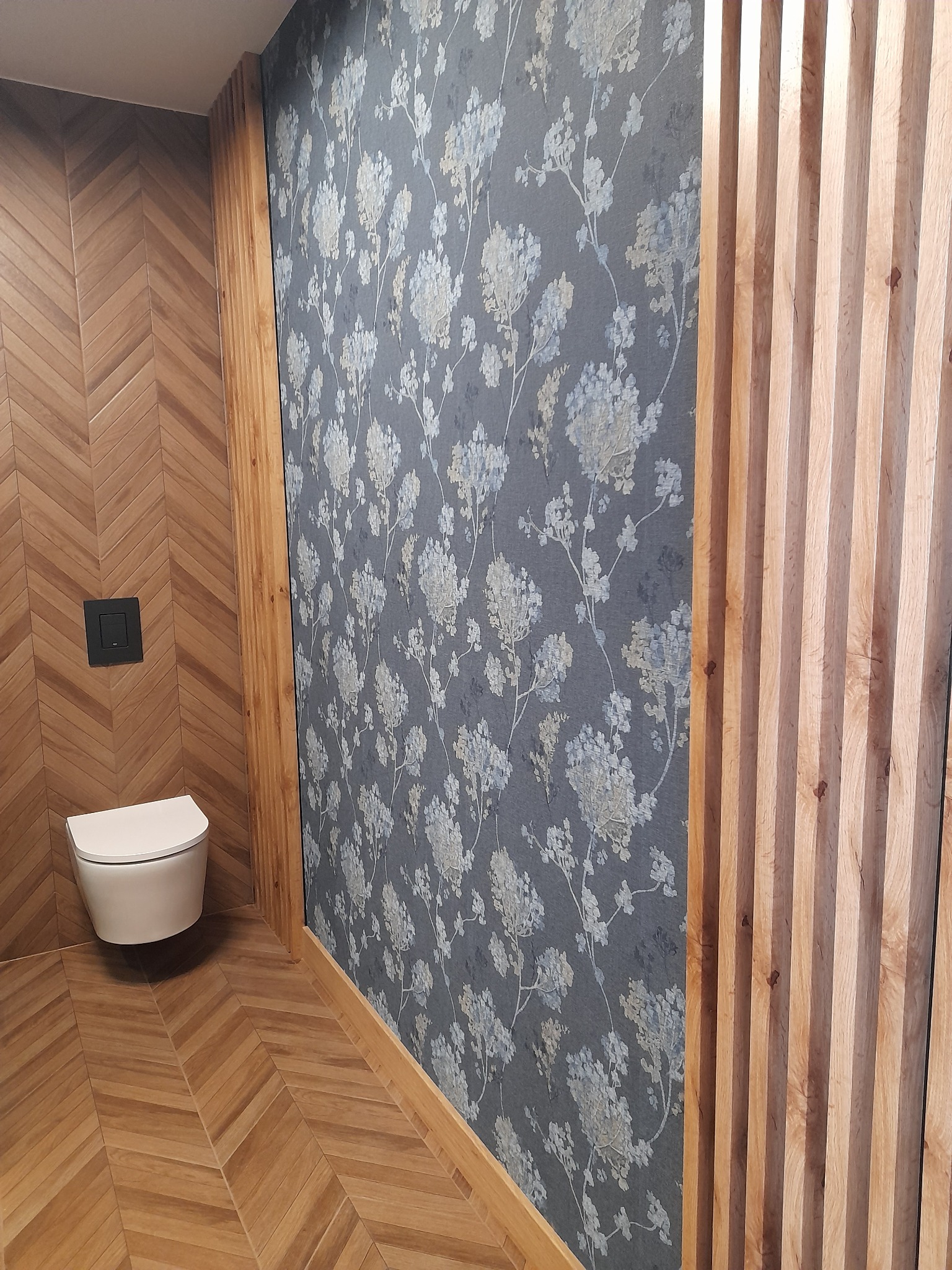 Nowoczesna łazienka z drewnianą okładziną ściany ułożoną w jodełkę, wisząca toaleta z czarnym przyciskiem spłukującym, ściana pokryta tapetą w kwiatowy wzór i dekoracyjne drewniane lamele.