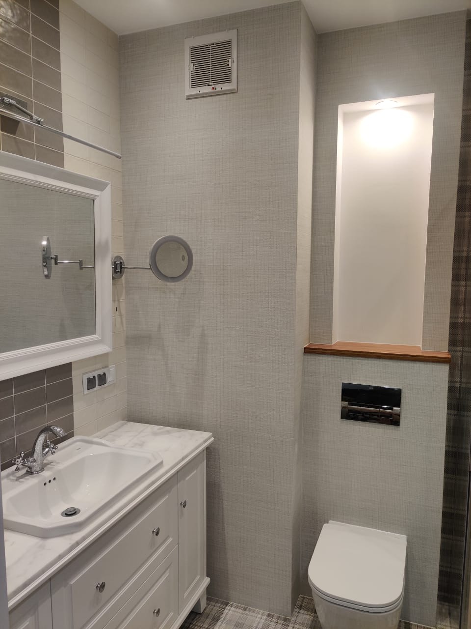 Jasna łazienka po remoncie: biała umywalka z szafką w stylu retro, toaleta pod zabudowaną półką z oświetleniem, lustro z oświetleniem punktowym na ścianie pokrytej tapetą o subtelnej fakturze.