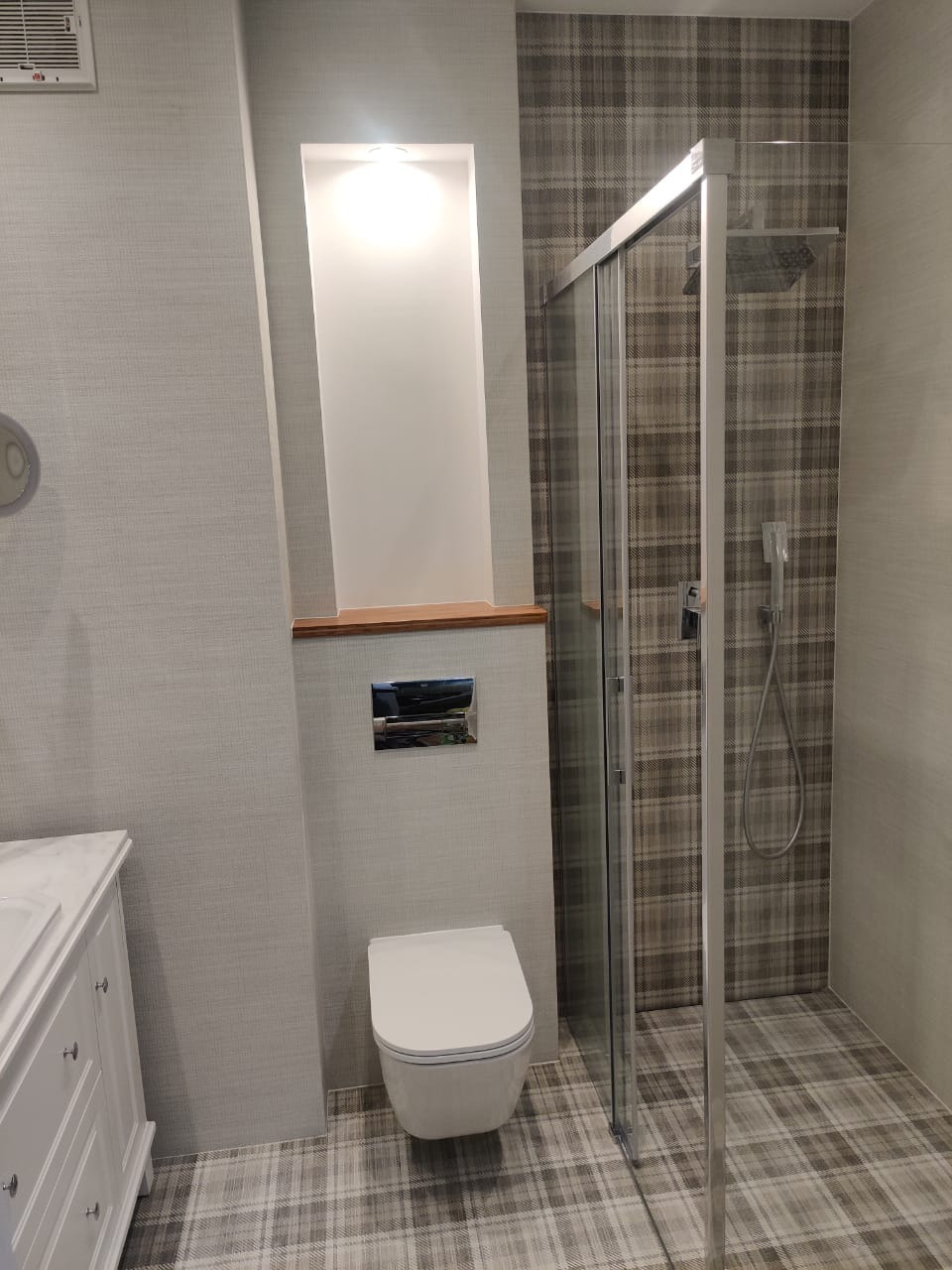 Nowoczesna łazienka z kabiną prysznicową i podwieszaną toaletą, wykończona płytkami w kratę, z wnęką oświetleniową i białą szafką pod umywalkę.