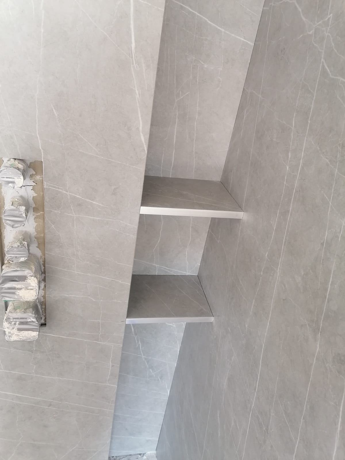 Ściana w łazience wyłożona szarymi płytkami imitującymi marmur z wnęką i półkami, widoczne pozostałości po usuniętych elementach na ścianie.