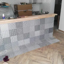 Montaż dekoracyjnych płytek ceramicznych w czarno-białe wzory na barze kuchennym, widoczne narzędzia i materiały budowlane, podłoga wyłożona panelami w jodełkę.