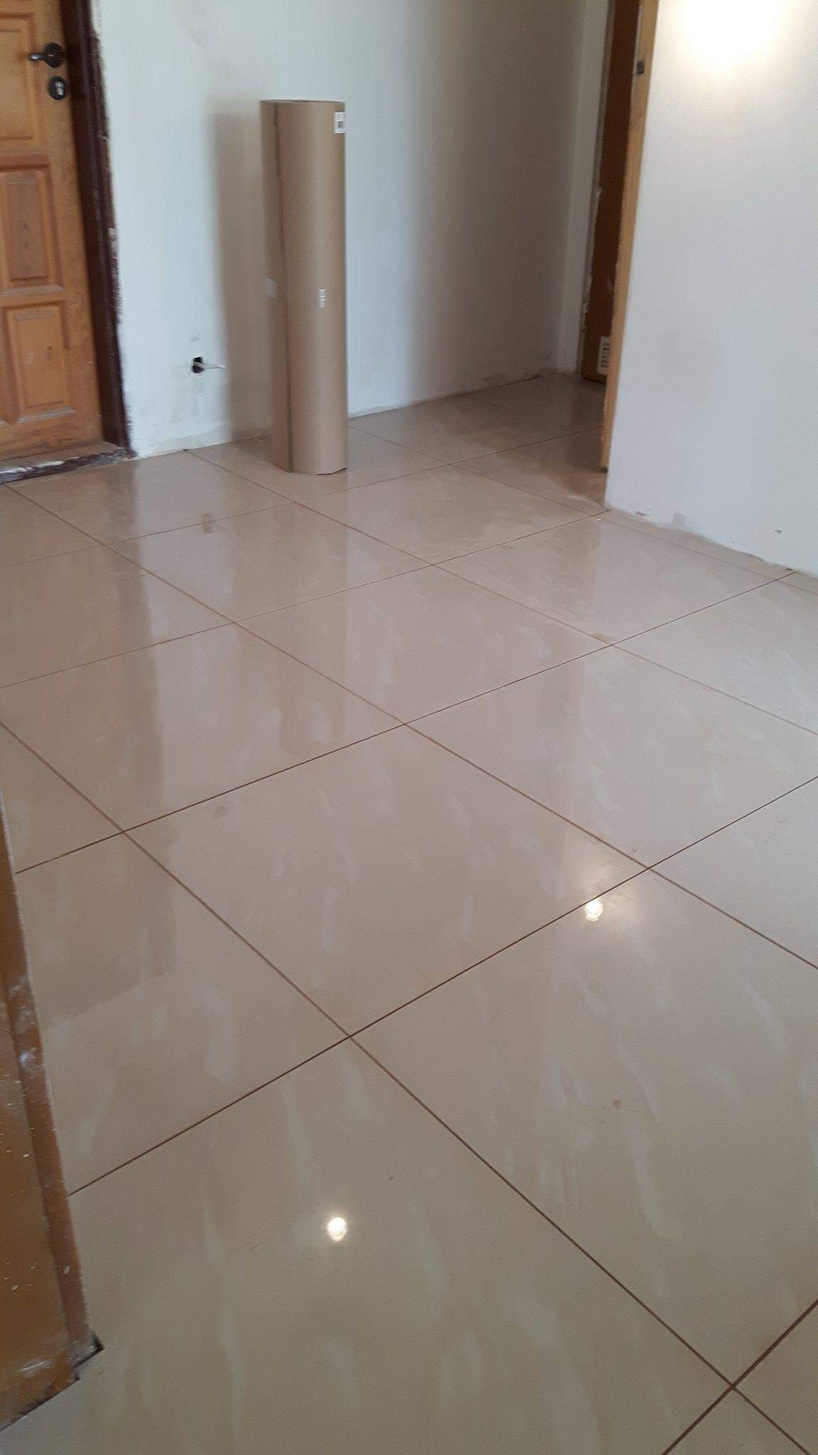 Błyszcząca, beżowa podłoga z płytek ceramicznych po ułożeniu, widoczna tubka z materiałem budowlanym i fragment drewnianych drzwi.