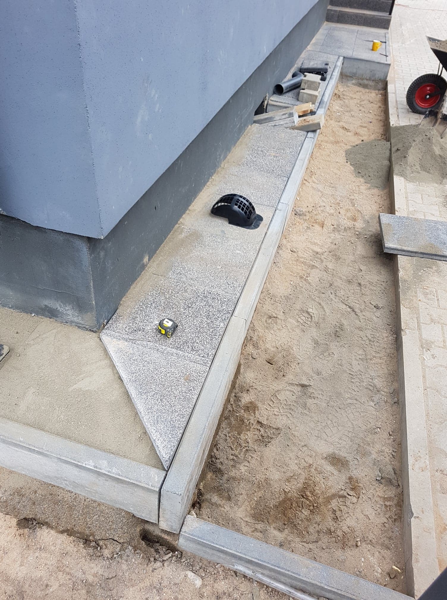 Układanie kostki granitowej wokół fundamentów budynku, widoczne elementy drenażu, ziemia i narzędzia pomiarowe.