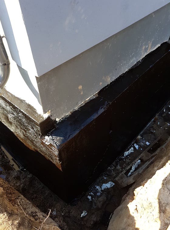 Hydroizolacja fundamentów budynku, widoczna czarna powłoka ochronna na styku ściany z gruntem, wykopane obok ziemne rowy.