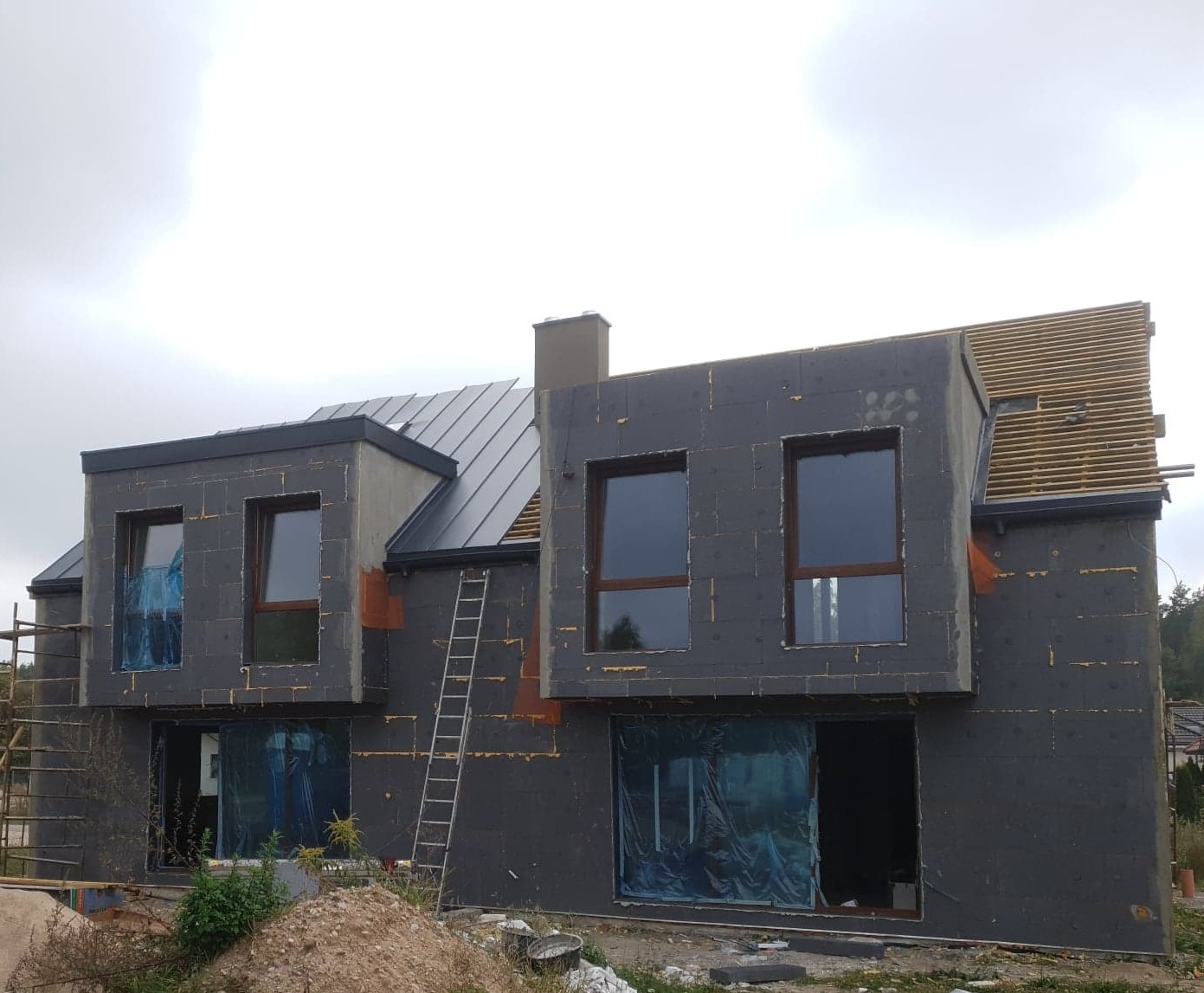 Dom w trakcie nakładania izolacji termicznej na elewację, widoczne płyty izolacyjne i rusztowanie, okna zabezpieczone folią, częściowo wykończony dach.