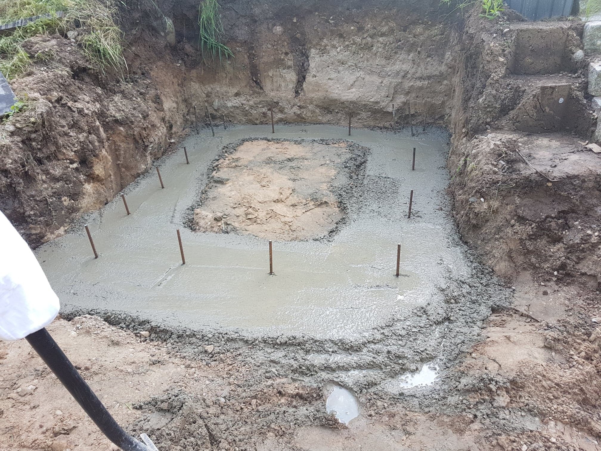Fundament w trakcie wylewania betonu, wystające pręty zbrojeniowe, widoczne ślady pracy łopaty, wykop w ziemi z roślinnością na górze.