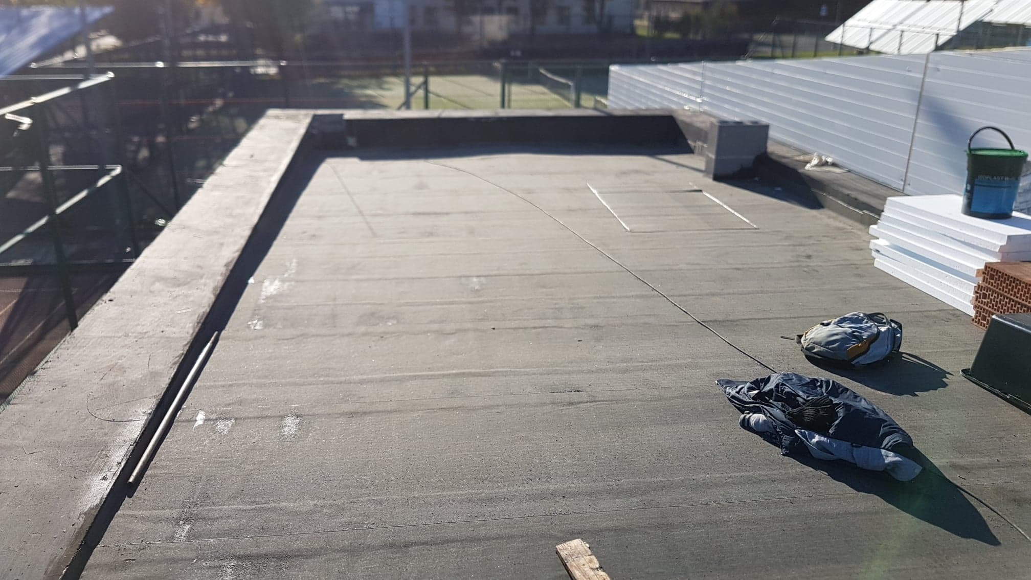 Płaski dach pokryty papą bitumiczną w trakcie prac: widoczny styropian do ocieplenia, wiadro z klejem, torba i ubranie robocze. W tle elementy konstrukcji dachu i ogrodzenie.