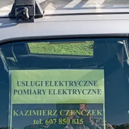 Ogłoszenie 'Usługi elektryczne, pomiary elektryczne, Kazimierz Czenczek, tel. 607 850 615' przyklejone do szyby samochodu z bagażnikiem dachowym i elementami instalacji elektrycznej.
